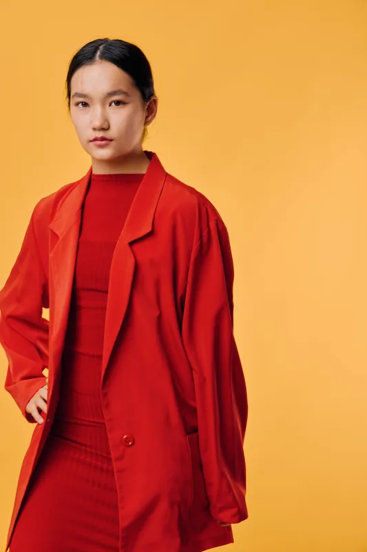 Young woman with black hair wearing red dress and oversized red blazer standing against yellow background with confident posture and calm facial expression.