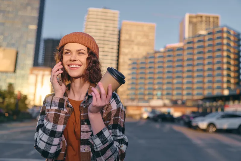 Woman on phone holding coffee in city street, smiling with candid expression and authenticity during golden hour glow; mindful living and emotional storytelling in urban lifestyle.