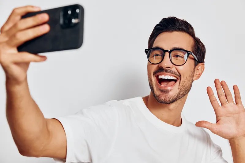 Smiling man with glasses taking a selfie using a smartphone, expressing joy and happiness, isolated on a plain light background, close-up shot.