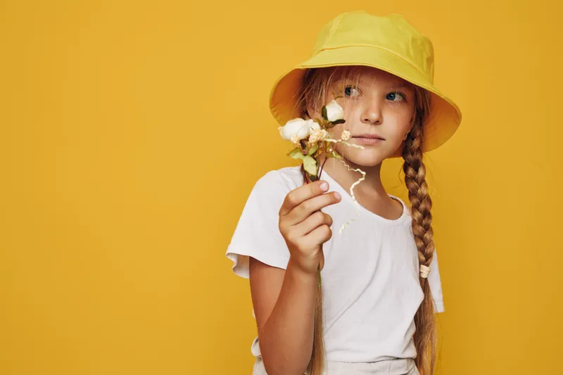 Young girl in a yellow hat holds dried flowers against a yellow background. She looks thoughtfully to the side in casual white clothing.