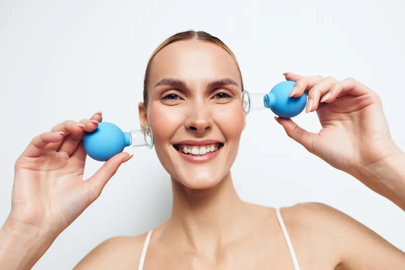 Young woman with clear skin holds two blue facial suction cups near her face smiling, demonstrating skincare routine and beauty treatment on white background.