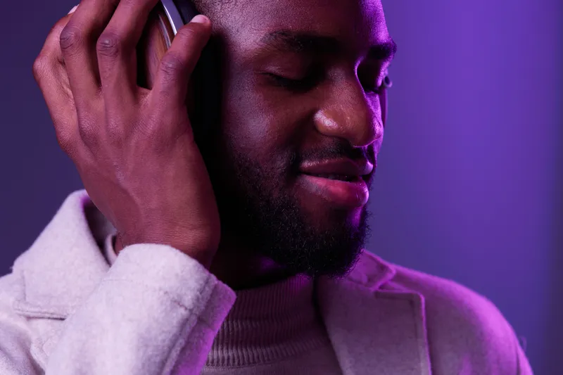 Happy young man enjoying music with headphones, wearing stylish clothes against a vibrant purple background, showcasing a joyful and relaxed atmosphere