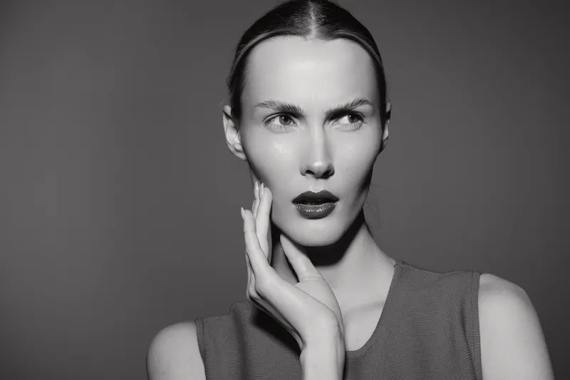 Young caucasian woman with light skin and slicked hair poses thoughtful with hand near face in studio with grey background.