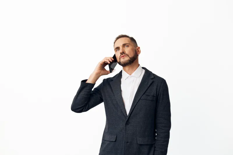 A professional man in a tailored suit stands against a clean white backdrop, conveying calm confidence, focus, and poised business demeanor during a phone call.