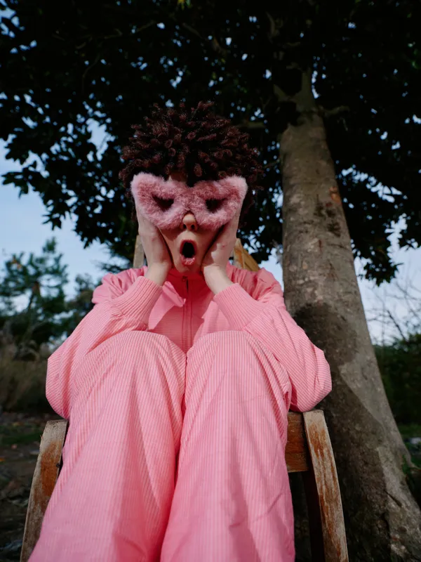 Woman in pink outfit sits on a wooden chair outdoors, hands on cheeks with mouth agape, conveying surprised expression under a leafy tree in a casual park scene with warm daylight