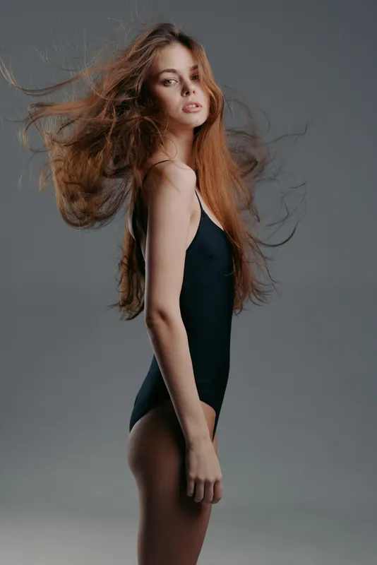 Young woman with long red hair wearing black bodysuit posing confidently with flowing hair in studio lighting
