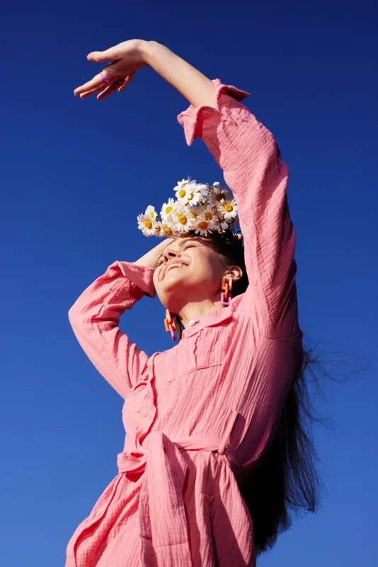 Joyful woman in a pink dress with flowers, expressing happiness against a clear blue sky, symbolizing summer and natural beauty