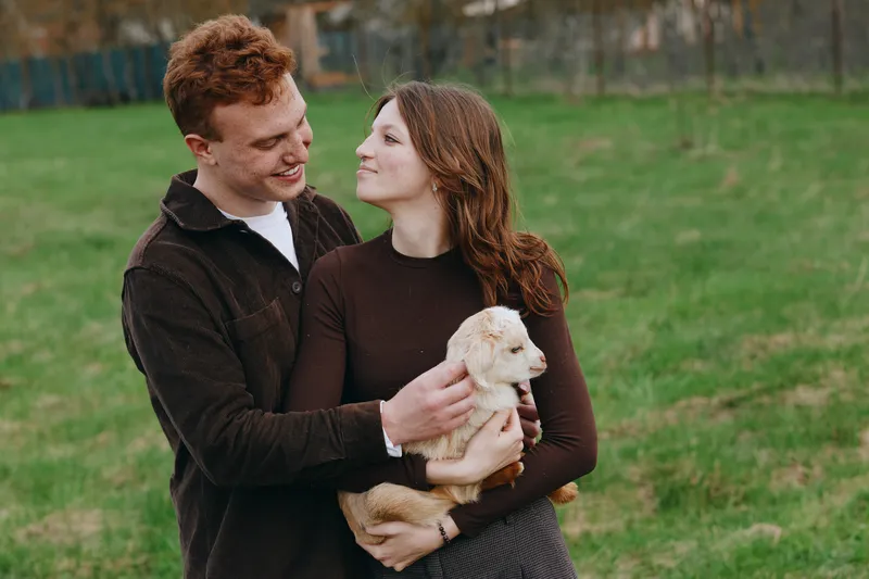 A young couple smiles in a sunlit park while holding a small puppy, showcasing affection and companionship in a casual lifestyle moment, warm and candid.