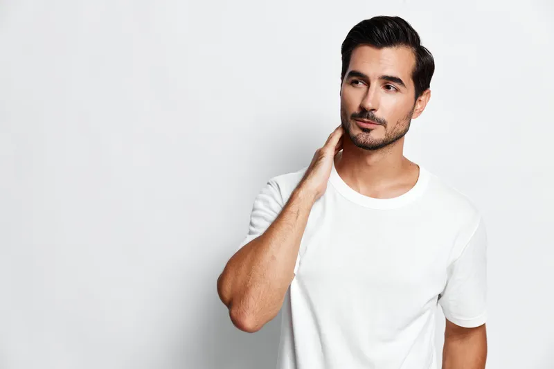 Handsome young man in a white t-shirt standing confidently against a solid white background, smiling softly while touching his neck. Lifestyle mood concept.
