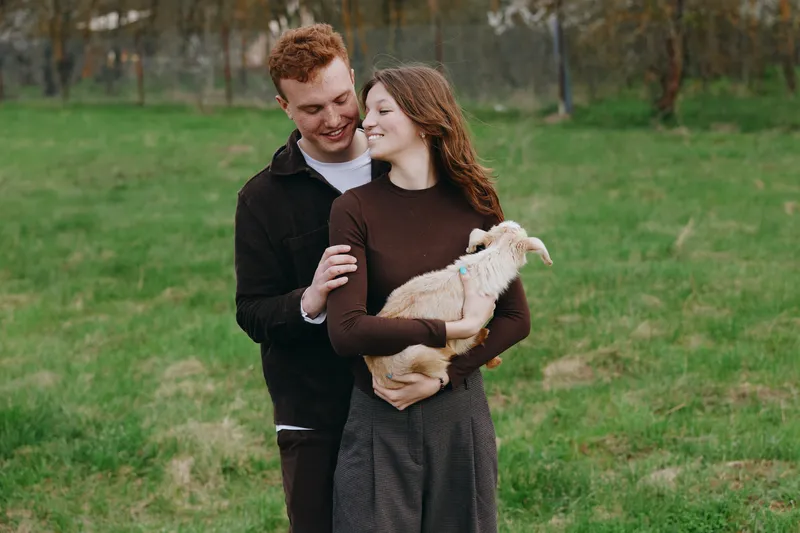 Cheerful couple outdoors, embracing gently, holding a goat kid in a meadow, warm natural light, relaxed mood, countryside setting, lush grass, intimate moment, lifestyle scene, candid photography
