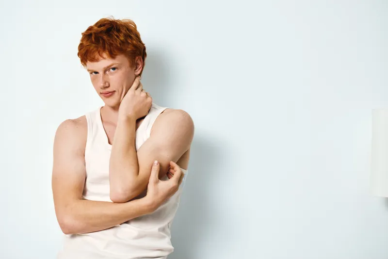 Young man with red hair wearing sleeveless white shirt standing against a plain wall looking confident and thoughtful with hand on neck in casual style portrait.
