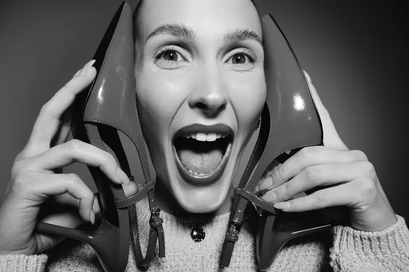 Cheerful Caucasian woman with fair skin holds red high heels near her face, smiling with open mouth. Concept for fashion, happiness, and lifestyle in black and white photography.