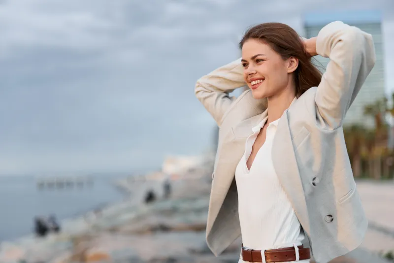 Confident businesswoman smiling outdoors in a stylish outfit, enjoying the coastal view while exuding professionalism and positivity.