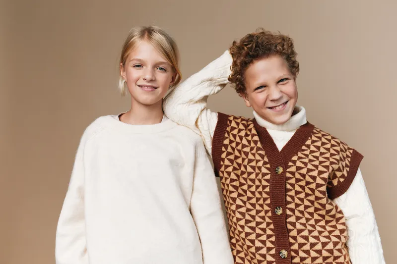 Two smiling children pose closely in cozy knit sweaters, showcasing warm textures, soft yarns, and friendly style against a neutral beige background.