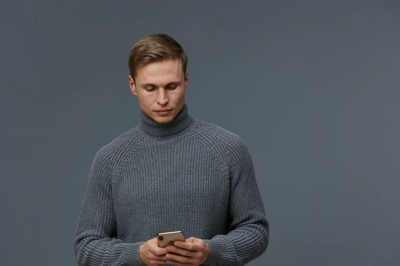 young caucasian man using smartphone wearing gray knitted sweater isolated on dark gray background casual winter look digital nomad concept