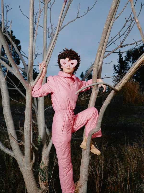 Person in pink jumpsuit climbs a tree with a heart shaped mask in a daytime outdoor setting, playful and surreal, fashion inspired, nature backdrop and bold color creating a whimsical portrait.