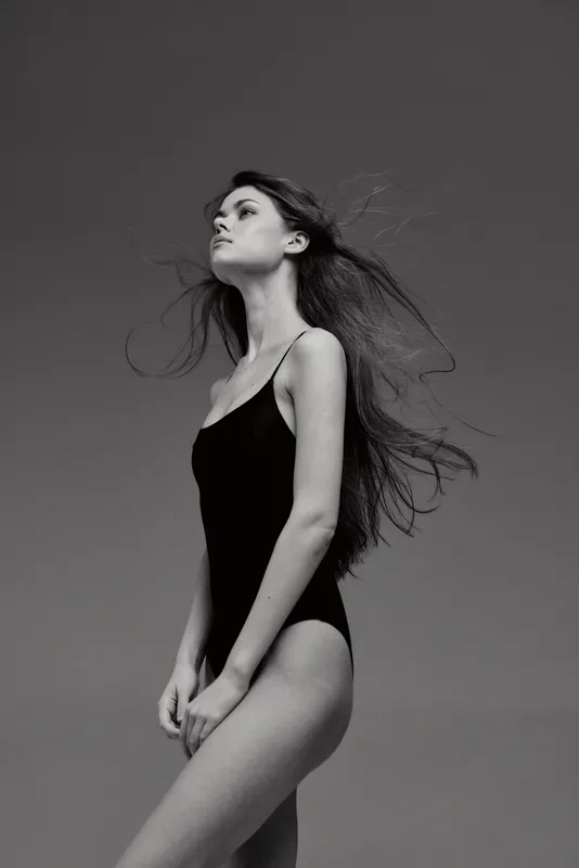 Black and white portrait of a slender woman in a black swimsuit with flowing hair and confident posture.