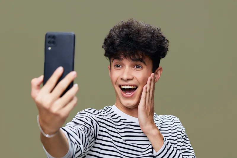 Young man taking a selfie with smartphone, expressing excitement and joy while wearing casual striped shirt against neutral background.