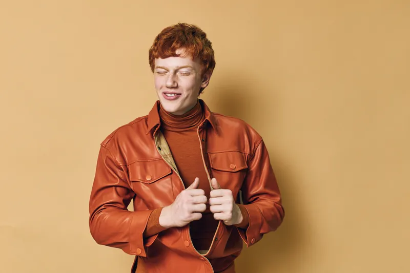 Young man with red hair and closed eyes wearing brown leather jacket and turtleneck sweater, posing confidently against beige background with relaxed expression.