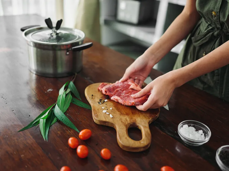 Женщина на кухне готовит ужин из мяса, овощей и специй на деревянном столе на фоне стильной кухни