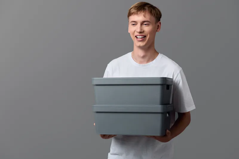 Young man in a white t shirt holding stacked gray plastic storage boxes, casual studio shot highlighting home organization, storage solution, and tidy living space for closet and moving needs