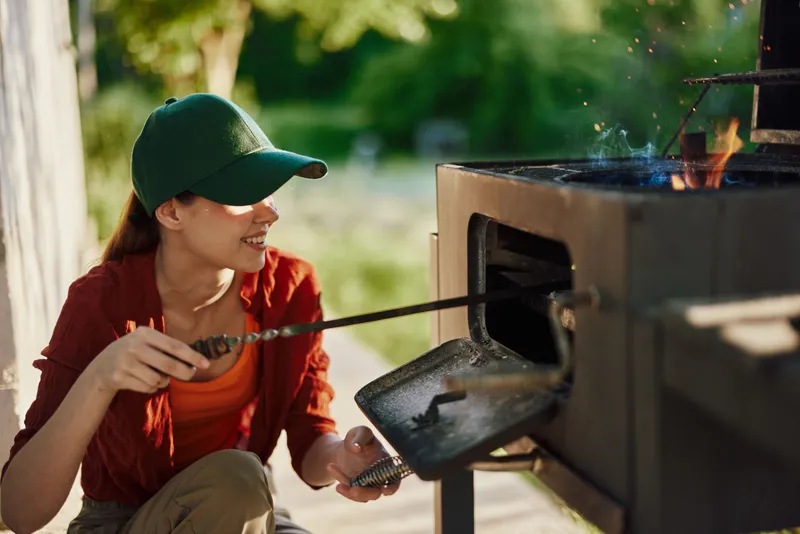 Young woman grilling outdoors, wearing a green cap and red shirt, smiling while cooking in a sunny garden setting
