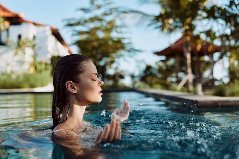 Young woman with wet hair relaxing in outdoor pool with closed eyes, enjoying calm moment in sunny tropical environment. Concept of wellness, lifestyle, and leisure time.