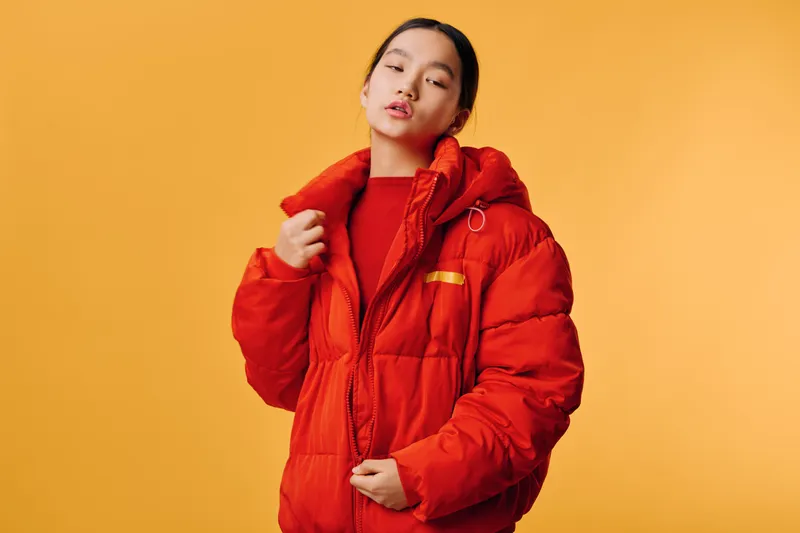 Young woman in red puffer jacket posing with eyes closed on orange background for fashion, style, winter clothing, and casual outerwear concept.