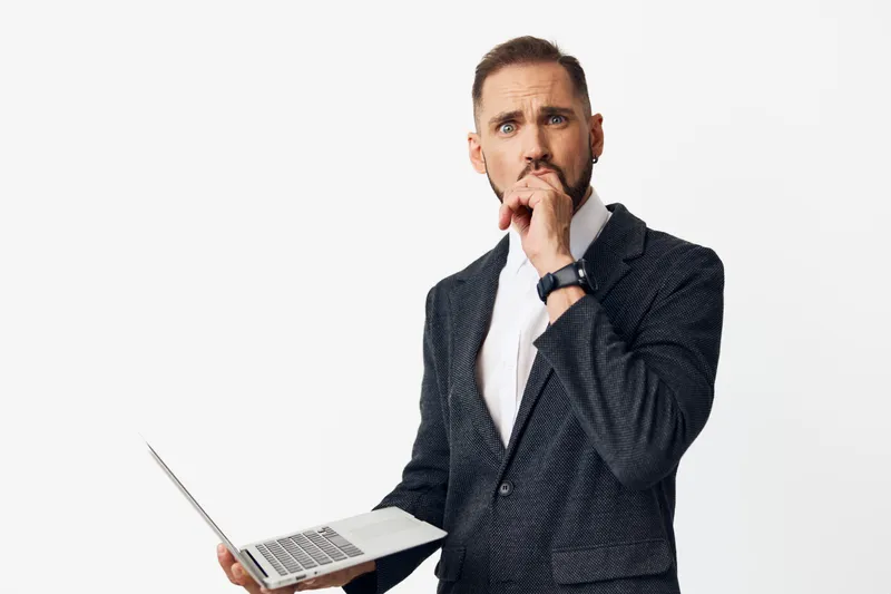 A focused business professional stands against a solid colored background, holding a laptop and studying the screen with a thoughtful expression, ready to solve challenges.