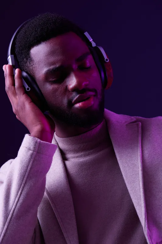 A young African man enjoys music with headphones, dressed in a stylish gray blazer and beige sweater, set against a vibrant purple background