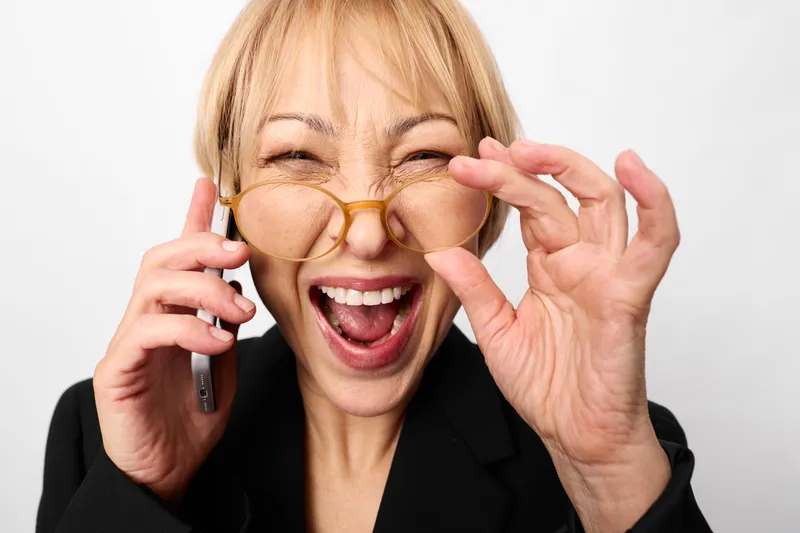 Cheerful woman with phone and glasses laughing in closeup, surprised expression, call in office, communication moment, white background, bright soft light today