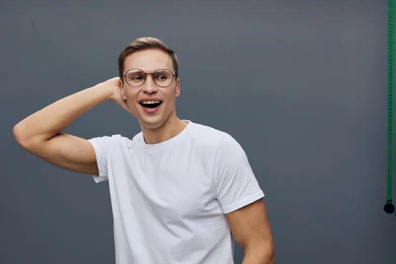 smiling man wearing glasses casually standing against a plain gray background, expressing joy and surprise, cheerful mood, modern style, casual clothing, portrait shot, studio setting