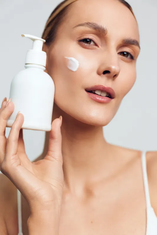 Close-up of a woman applying white face cream on cheek holding a lotion bottle. Concept of skincare, beauty, wellness, and daily self-care routine with soft natural lighting.