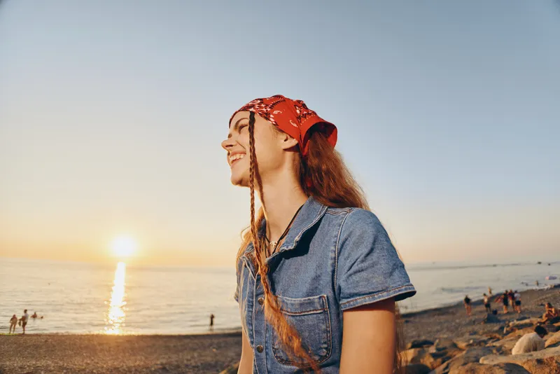 Lifestyle woman wearing modern boho-western denim street style and red bandana, smiling joyfully at sunset on the beach with film color tones.