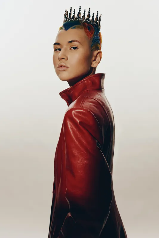 Young woman with colorful hair and a black crown wearing a red leather jacket, posing confidently against a light background in a modern studio setting.