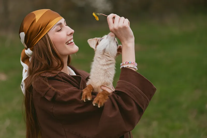 Smiling woman in a brown outfit and headscarf plays with a goat kid in a sunny park, sharing a joyful moment of connection with nature and animals. Warm light highlights the joyful moment.