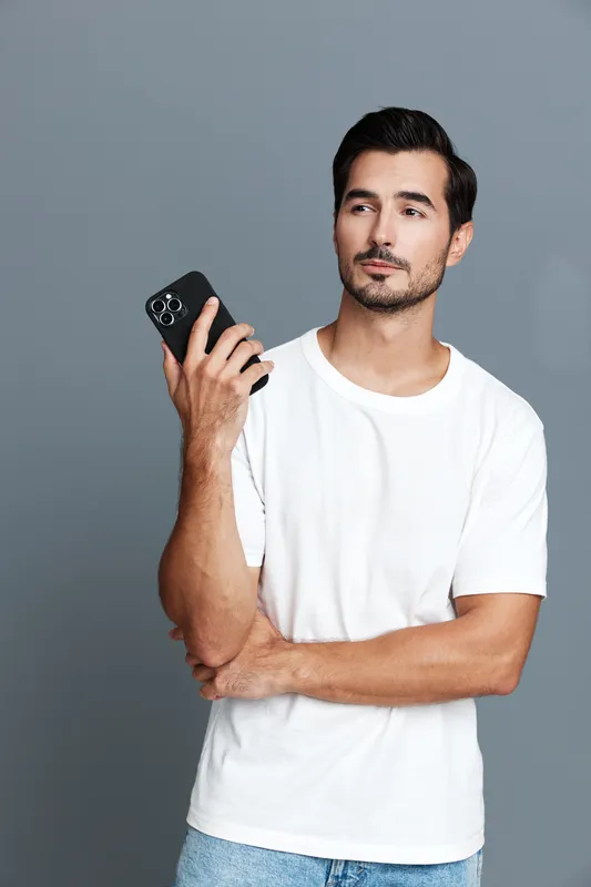 Casual thoughtful young man with smartphone in hand looking up and pondering, dressed in a white t-shirt and light jeans, against solid grey background. Lifestyle concept