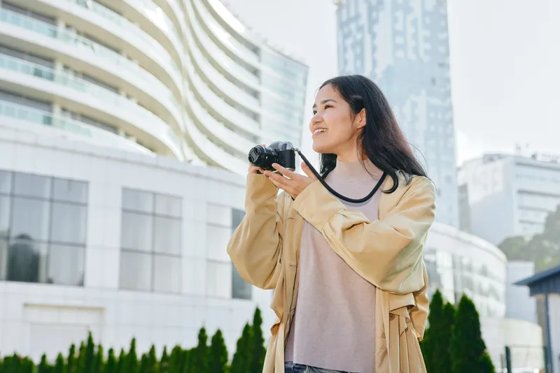 Woman with a camera in an outdoors urban setting, smiling while capturing city architecture, daylight lighting, fashionable jacket, candid travel photography moment in the city