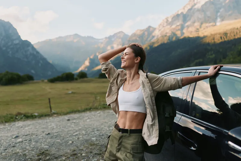 Woman outdoor lifestyle portrait beside a car in a dramatic mountain landscape, casual clothing, fresh air energy, relaxed smile, enjoying nature during a scenic travel moment