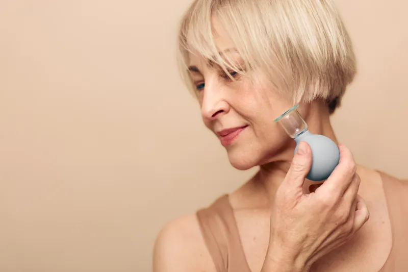 Mature woman with short blonde hair gently applies facial cleansing brush on beige background. Concept of skincare, beauty routine, personal care, and healthy skin for older adults.