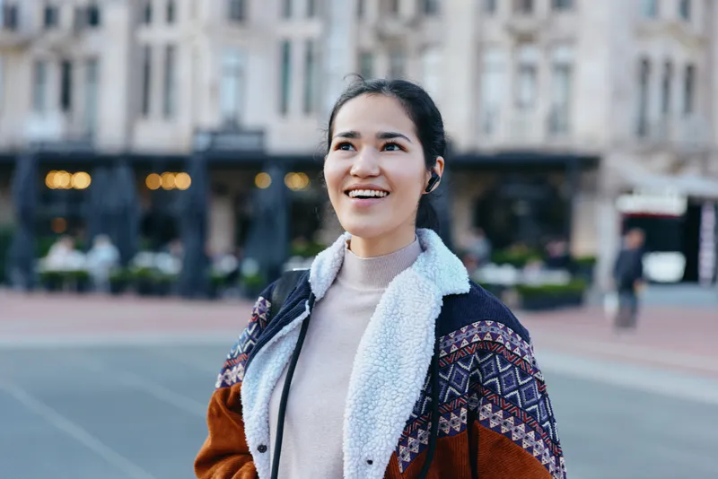 Woman walking in a city street, smiling outdoors in a cozy sweater and colorful jacket, urban backdrop of buildings, candid moment, casual fashion, confident mood, everyday life, streetstyle energy