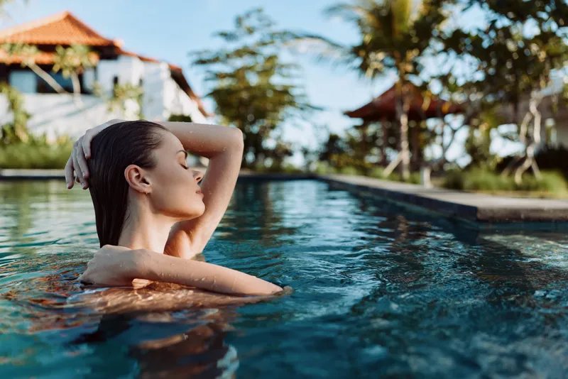Woman in an outdoor pool with wet hair enjoying a tranquil moment. Sunlit blue water glitters, conveying lifestyle, wellness, and self care in a bright resort setting.