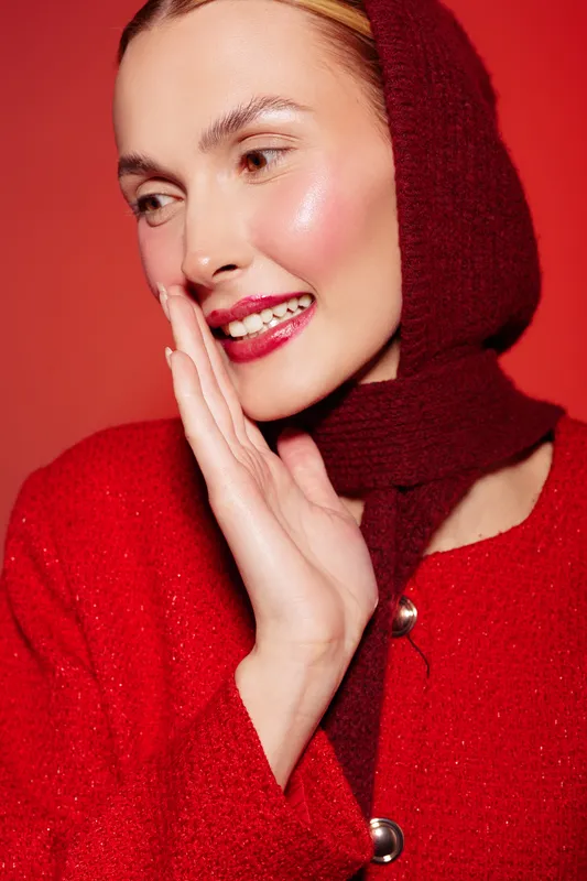 Smiling woman in red woolen hood and coat touches face with hand, looking aside on red background. Concept of winter fashion, beauty, and cozy seasonal style.