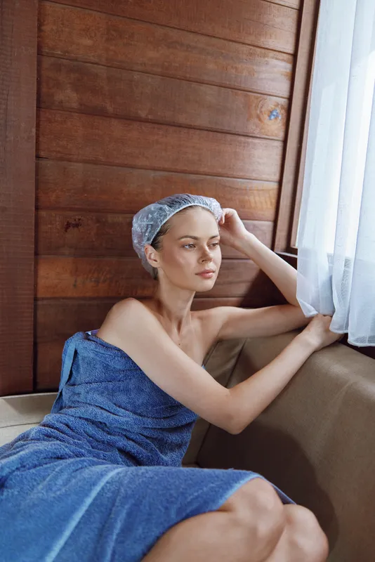 Young woman with thoughtful expression wrapped in blue towel and wearing shower cap, sitting near window with wooden wall background, natural light softly illuminating her face.