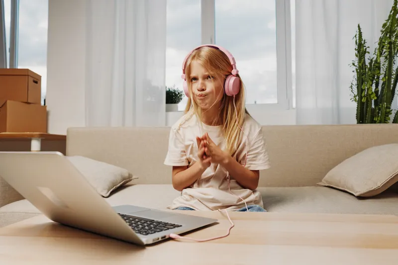 Cute little girl with long blonde hair wearing pink headphones enjoys music at home. She sits on a beige couch near a laptop in a bright living room with plants and cardboard boxes.