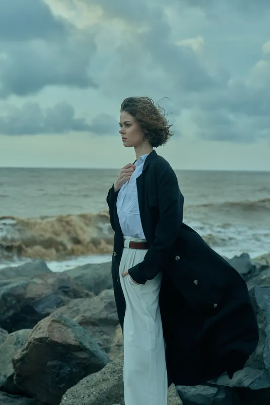 Windy coastline, young woman in white pants and black coat, standing with ocean breeze, contemplative mood amid rocks.