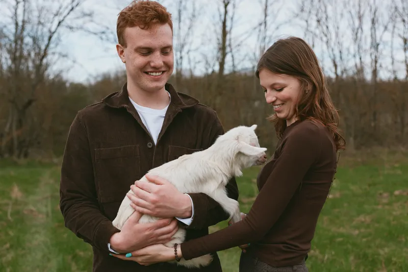 Cheerful couple outdoors, smiling while cradling a goat kid, sunlit meadow scene, relaxed posture, natural colors, candid lifestyle moment, warm interaction in a peaceful field setting
