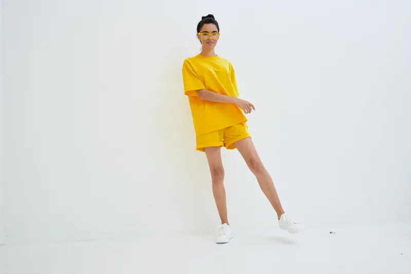 young woman wearing yellow casual outfit and matching sunglasses standing against white wall, minimalistic style, bright cheerful summer look