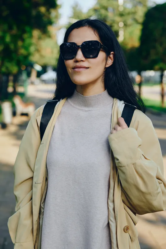 Woman in casual fashion wears sunglasses and a beige coat in a park, relaxed street style, confident pose, soft sunlight and green backdrop for a lifestyle shot.