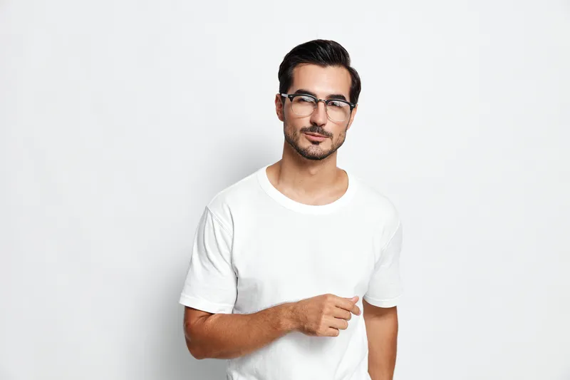 Casual young man in white t-shirt with stylish glasses posing confidently, smiling at the camera against a minimalistic solid background. Modern lifestyle concept.
